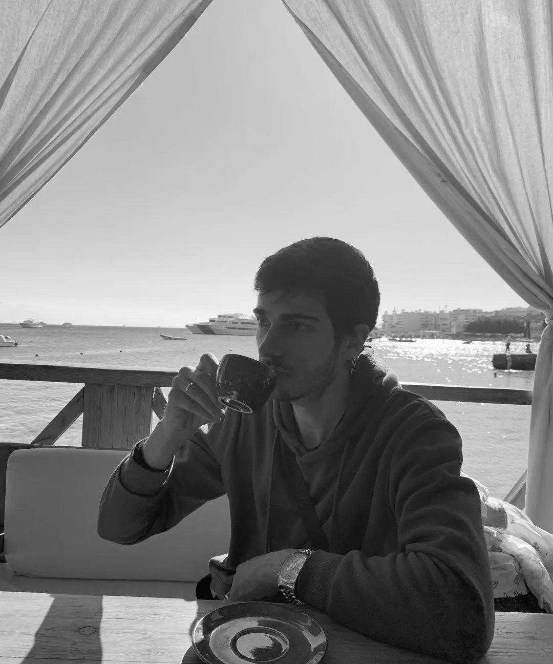 Sayid Bilal seated at a table by open water, drinking from a dark cup.