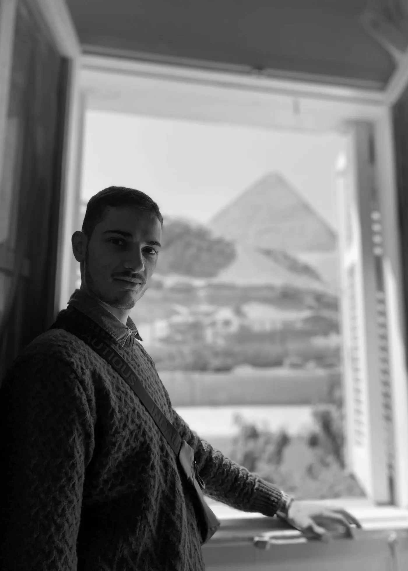Sayid Bilal at an open window, looking back to camera, a pyramid in the daylight beyond.
