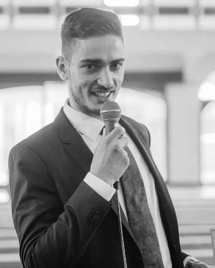 Sayid Bilal in a suit, speaking into a handheld microphone in a bright hall.